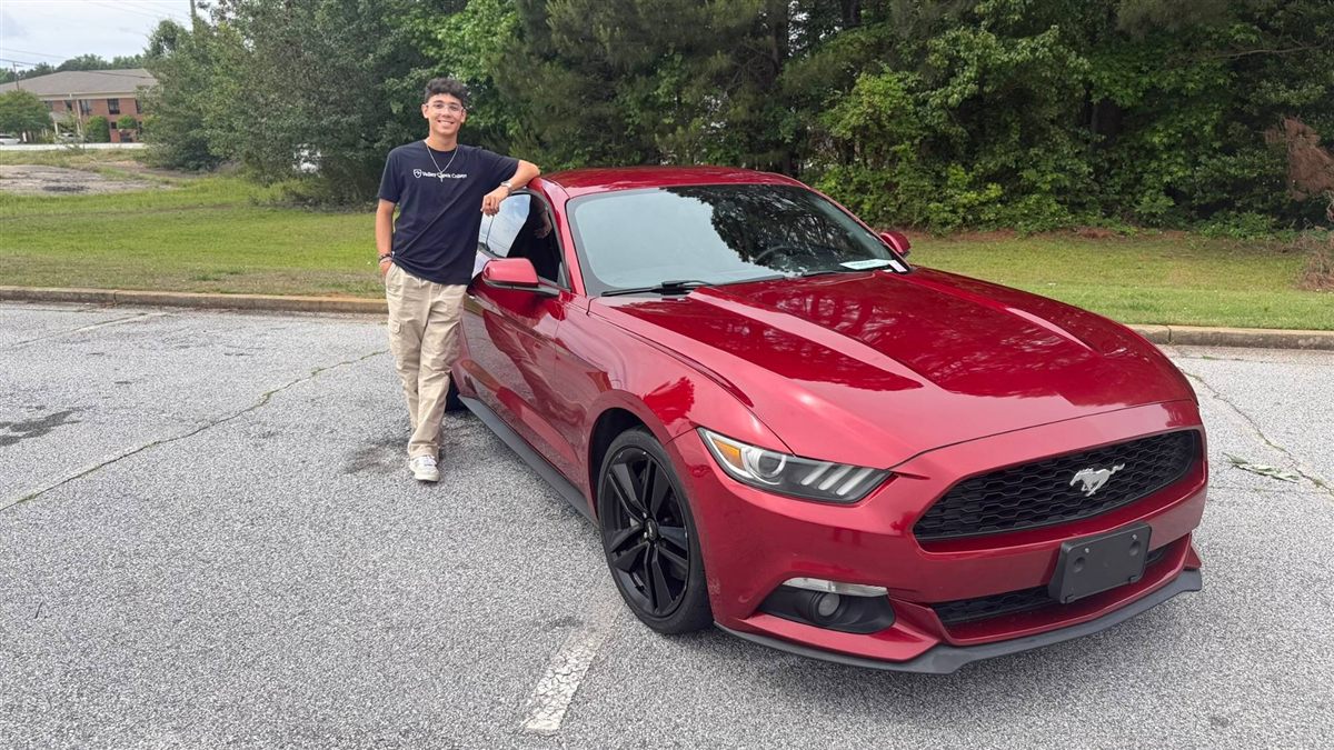Nathan smiling and leaning on his Ruby Red Ford Mustang in a parking lot